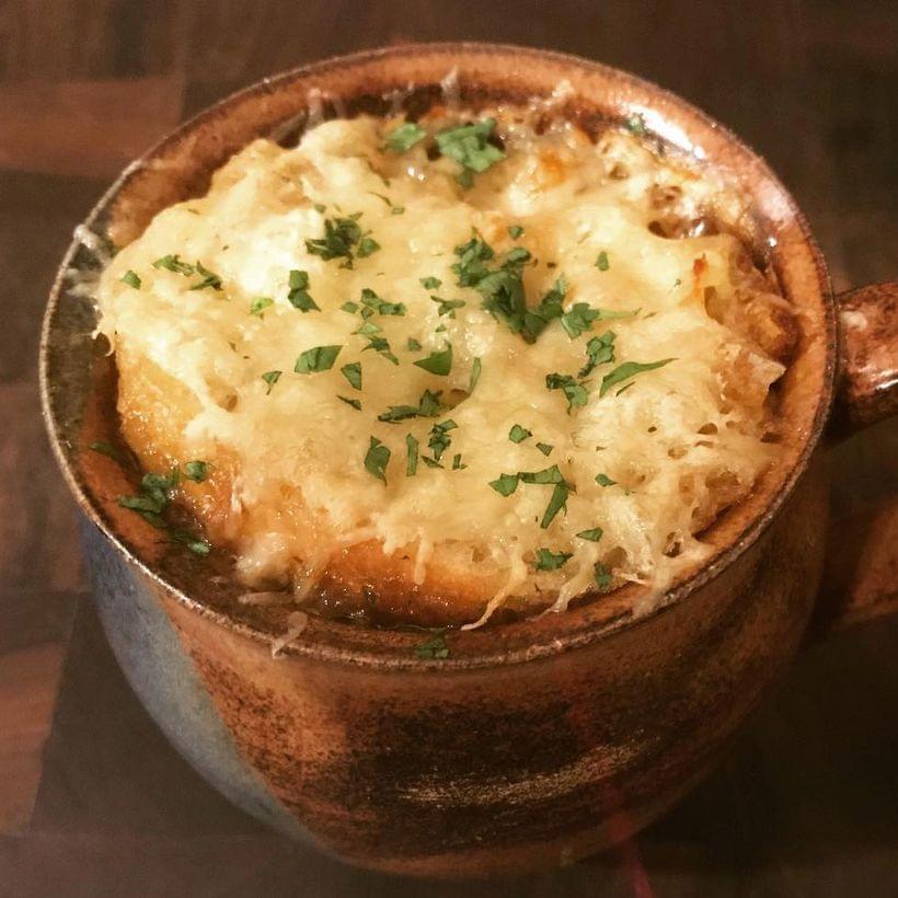 An earthenware mug of french onion soup, topped with a sourdough crouton, melted cheese, and a sprinkling of finely chopped parsley