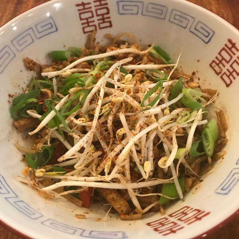 A bowl of dark brown rice noodles, topped with raw bean sprouts and scallion greens.