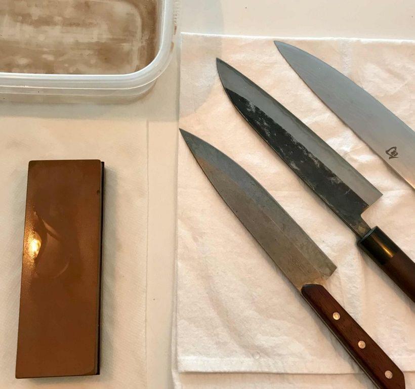 Three chef’s knives side-by-side on a cloth rag, alongside a dampened sharpening stone and a bowl of water.